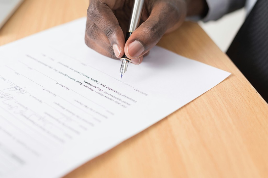 A close-up of a hand holding a pen, signing a document placed on a wooden desk.